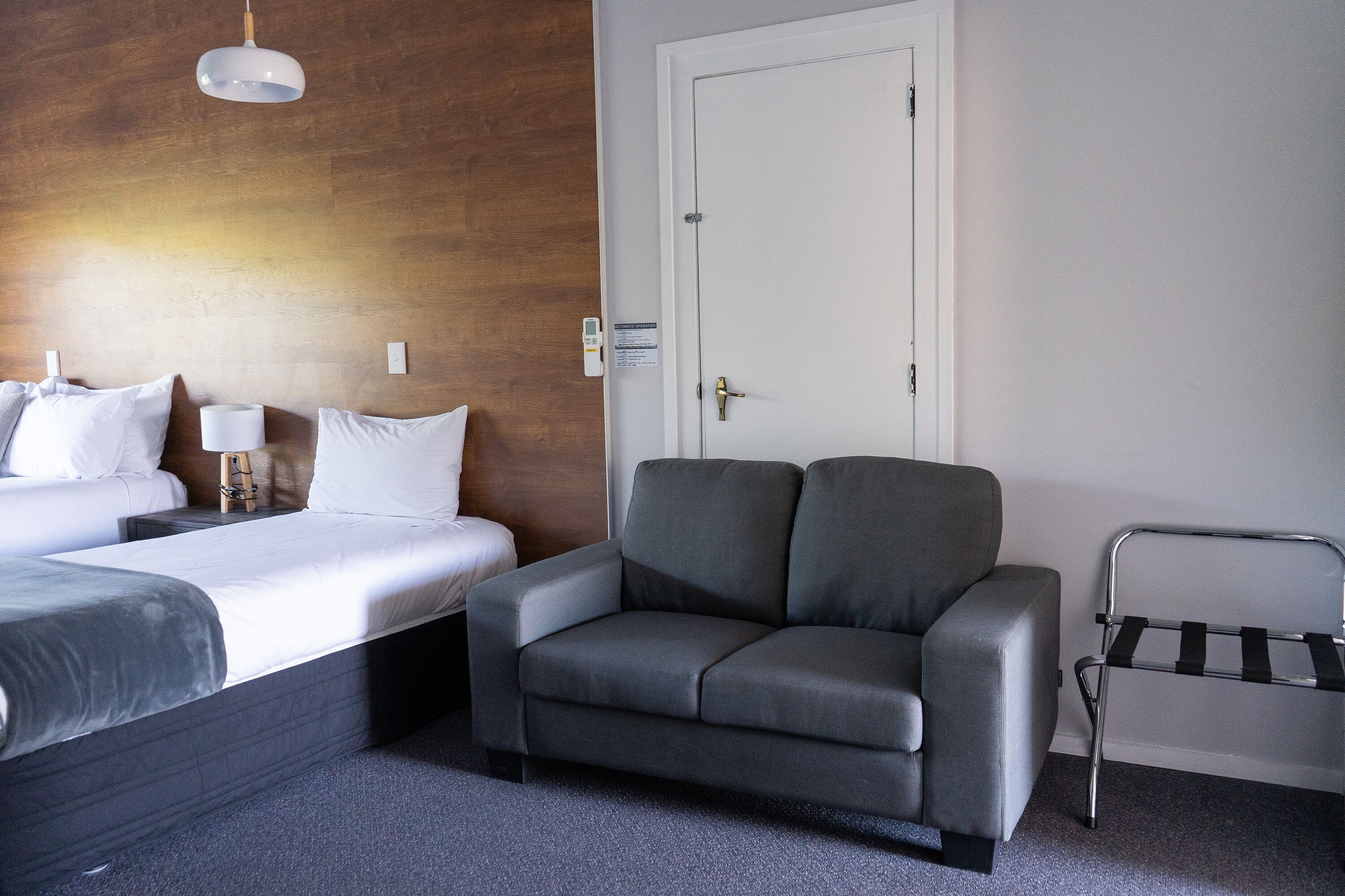 Habitación de hotel con una cama, un sofá gris, una mesita de noche con una lámpara, un perchero y una pared de madera.