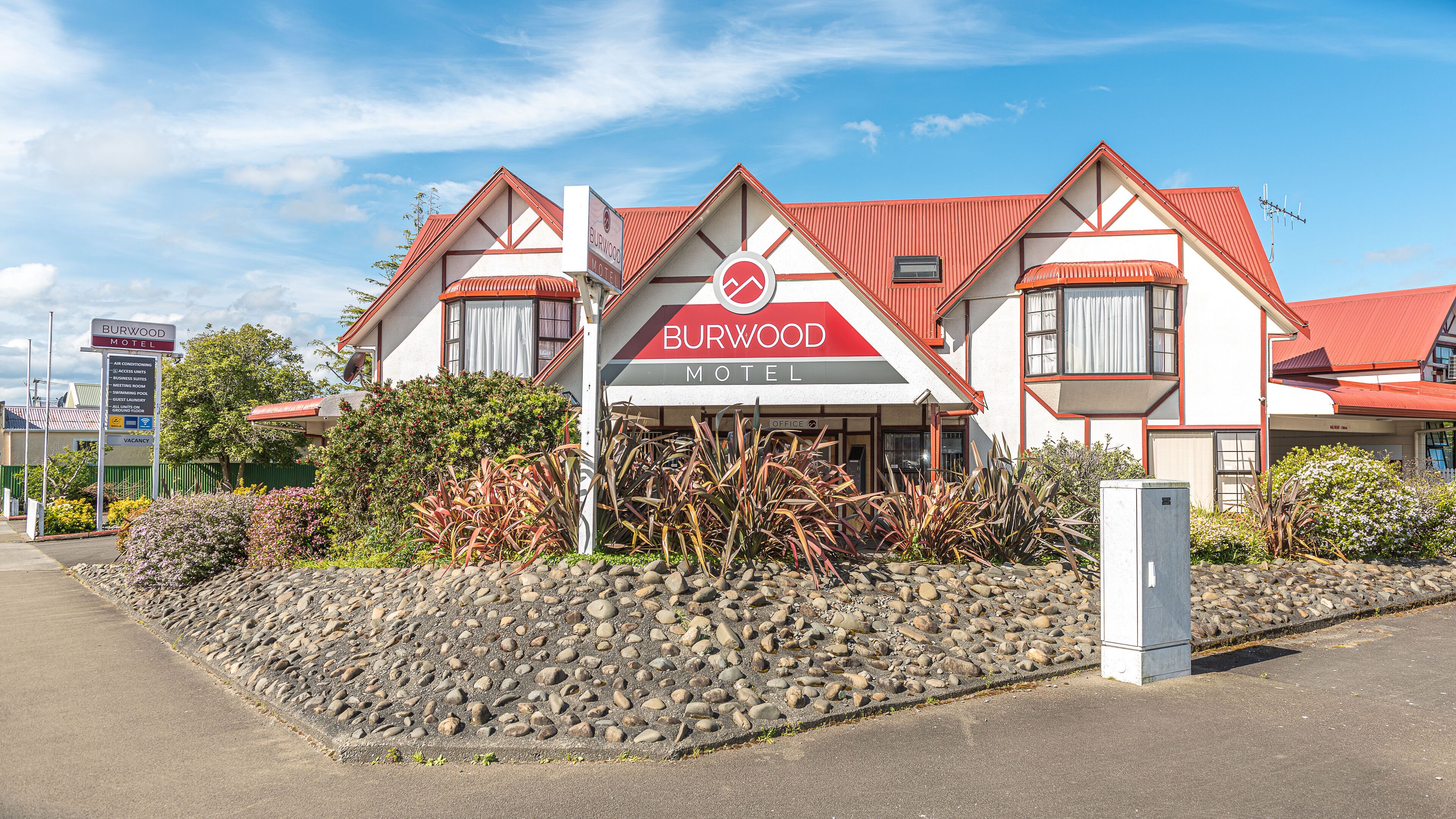 Un motel à la façade rouge et blanche, avec un panneau indicateur et un jardin aménagé devant, agrémenté de rochers et de plantes.