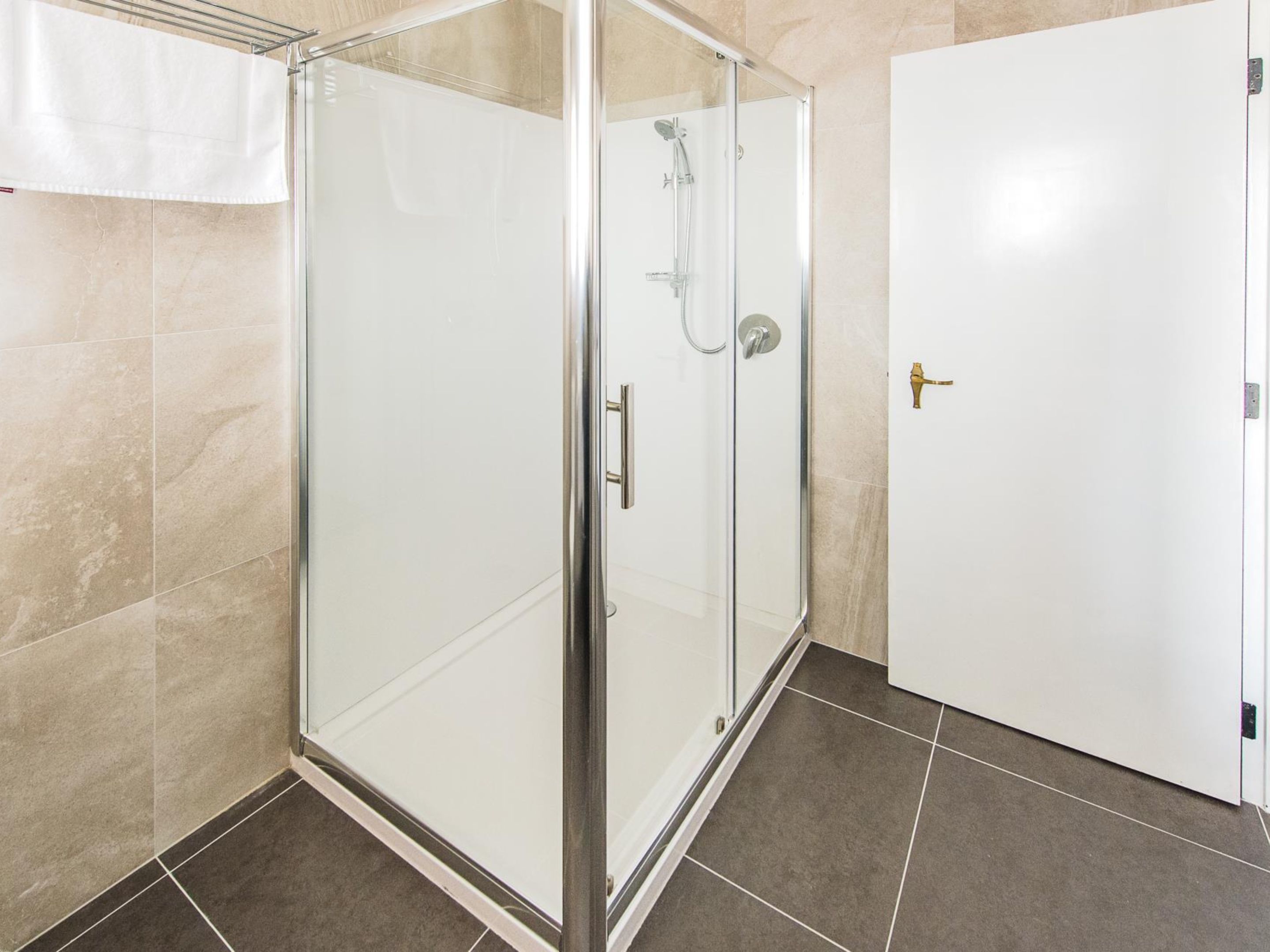 A glass-enclosed shower with a handheld showerhead, a door with a brass handle, and tiled flooring.
