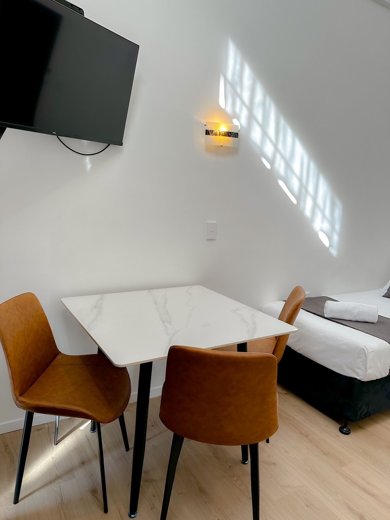 A modern room with a white table, two brown chairs, a bed, and a TV mounted on the wall.