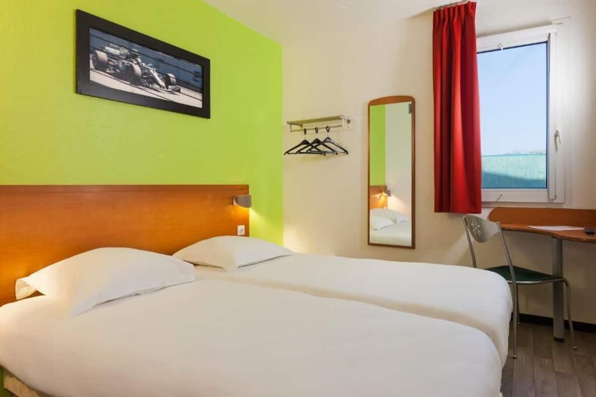 Une chambre d’hôtel avec deux lits, un bureau, une chaise, une fenêtre avec un rideau rouge, un miroir et un tableau encadré au mur.