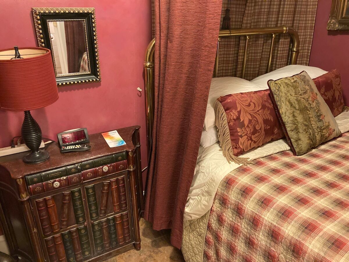 A bedroom with a bed, a nightstand with books, a lamp, and a mirror.