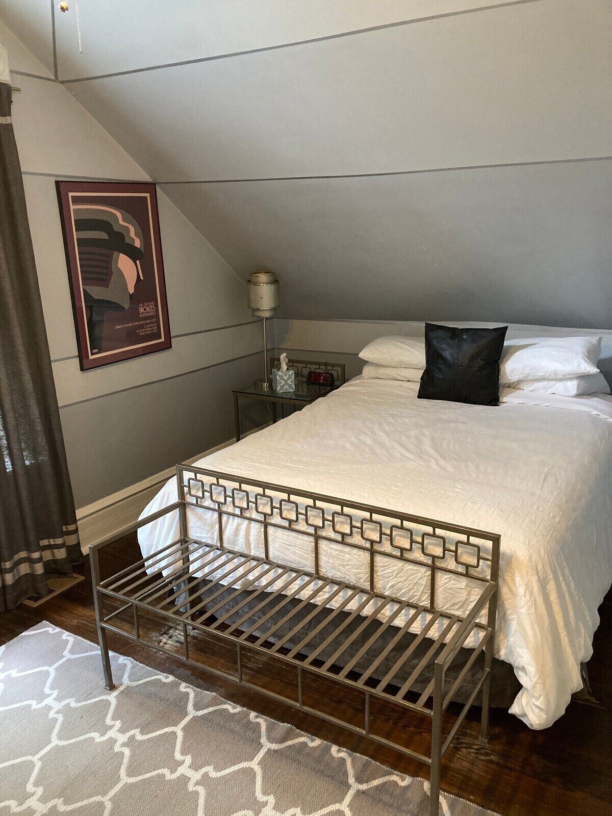 Un dormitorio con cama, mesita de noche, lámpara, cuadro enmarcado y ventana con cortinas.