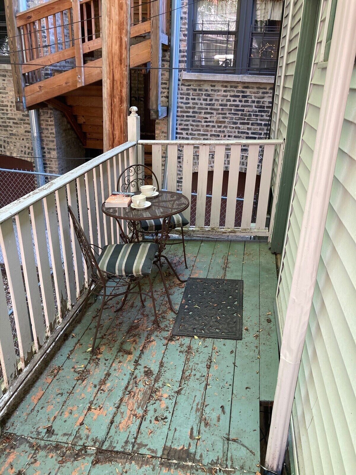 A small balcony with a table and chairs, a metal railing, and a view of a brick building.