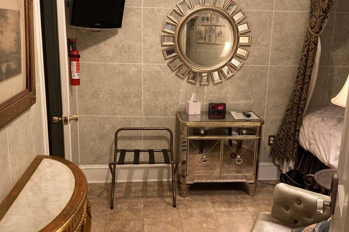 A bathroom with a vanity, a mirror, a television mounted on the wall, a small table with toiletries, and a chair.
