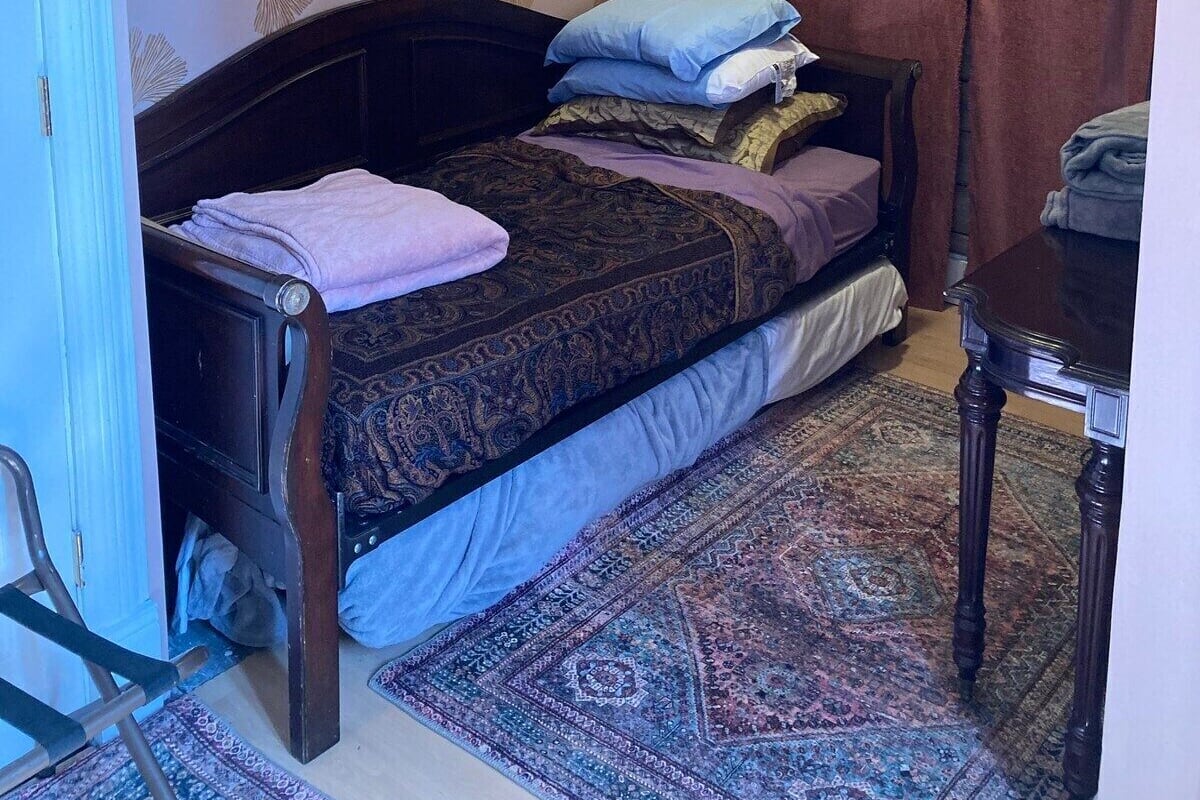 A bedroom with a bed, a wooden chair, a small table, a patterned rug, and a curtain.