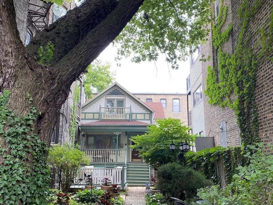 A narrow stone pathway leading to a house with a green porch and a large tree overhanging the path.