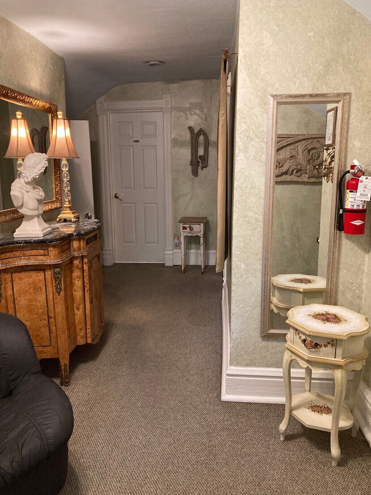 Una habitación con un aparador de madera, una escultura de busto y un espejo con diseño floral. Hay una puerta blanca y una pequeña mesa blanca con un extintor rojo en la pared.
