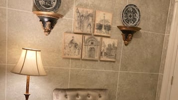 Un sillón con volante, un cojín estampado, una lámpara, y platos decorativos y cuadros colgados en una habitación con paredes de azulejos.