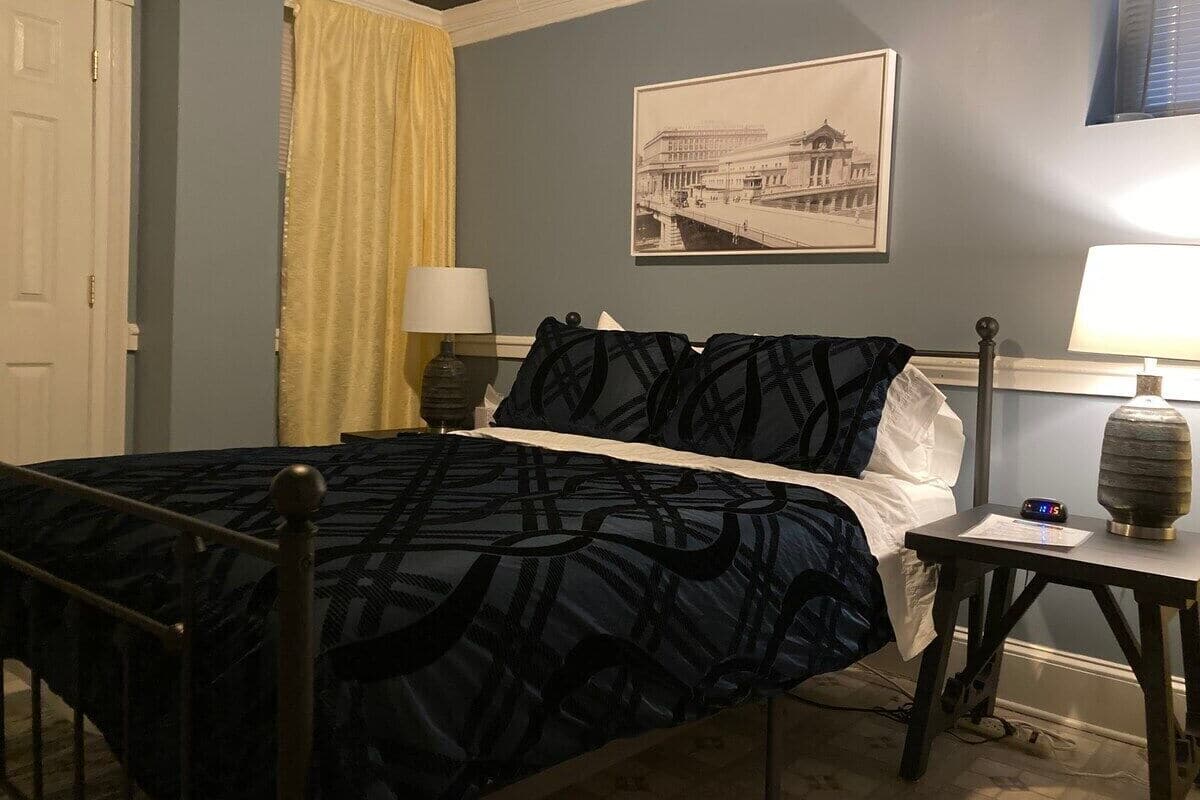 A neatly made bed with a patterned bedspread, two bedside tables with lamps, a framed picture on the wall, and a window with blinds.