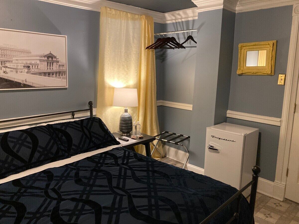 A hotel room with a bed, a small table with a lamp, a minibar, a coat rack, and a framed picture on the wall.