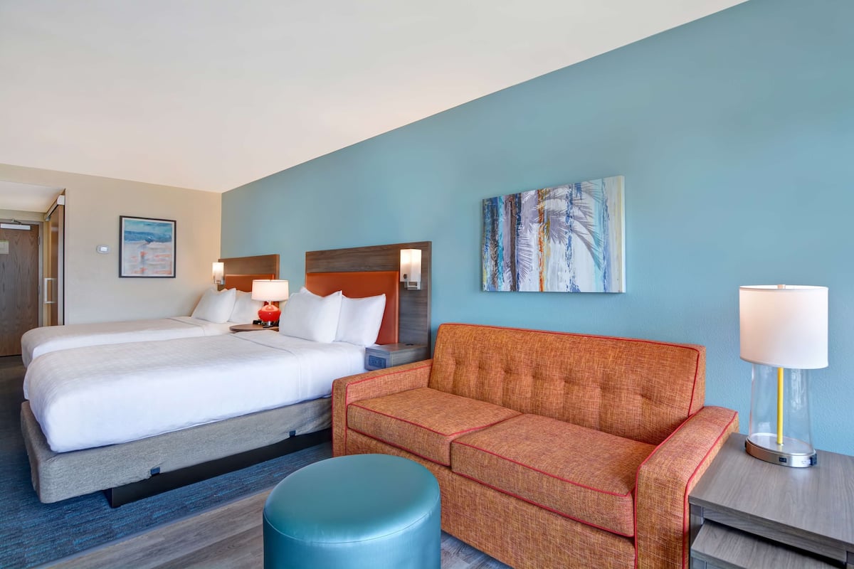 Une chambre d’hôtel avec deux lits, un canapé, un pouf rond et un tableau au mur.