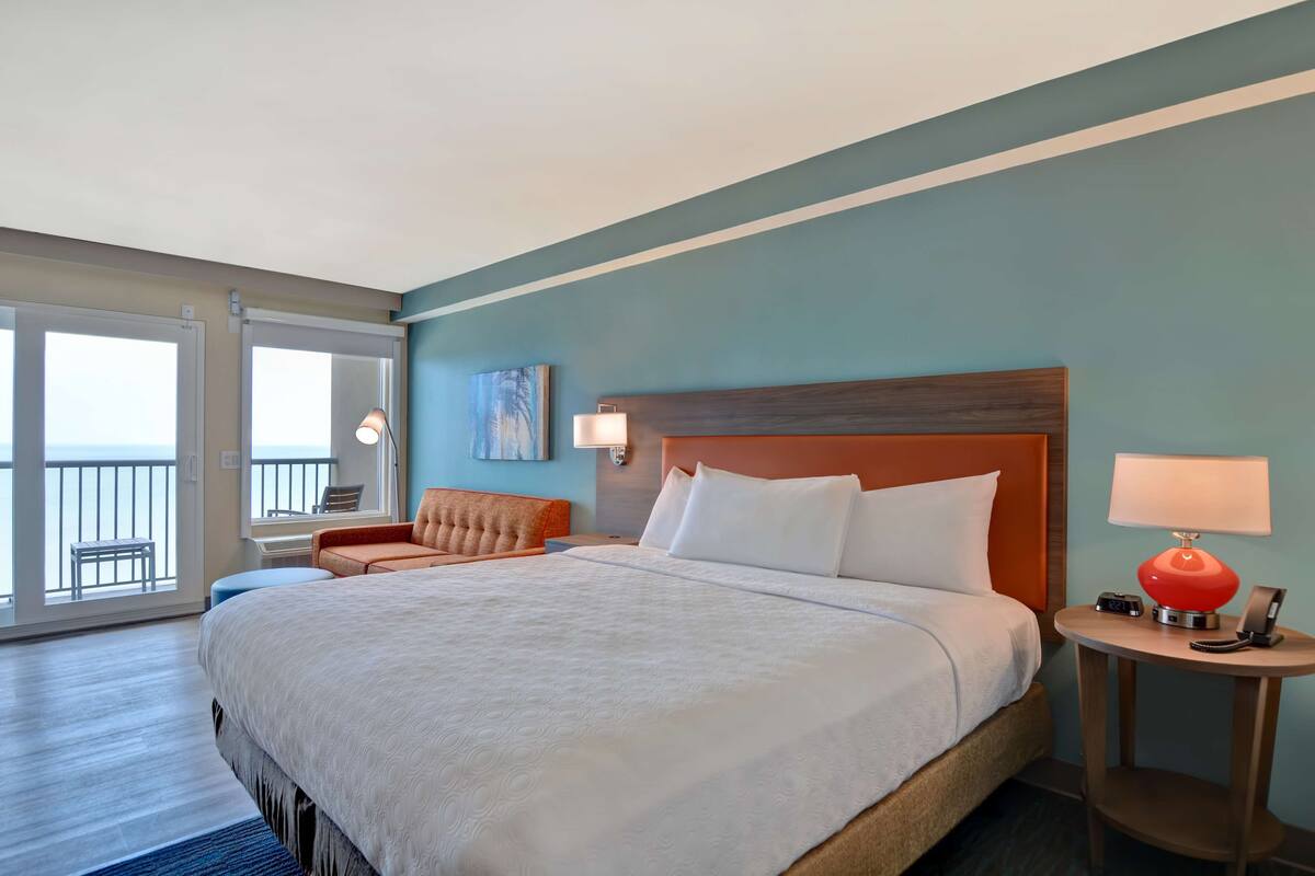 Une chambre d’hôtel avec un grand lit, un petit canapé, une table de chevet avec une lampe et une vue sur l’océan grâce à de grandes fenêtres.