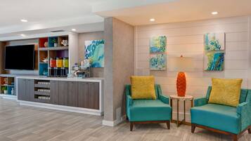 A modern living room with teal armchairs, a flat-screen TV, and a kitchen area with a built-in fridge and shelves.