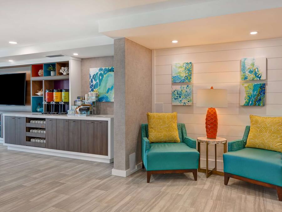 A modern living room with teal armchairs, a flat-screen TV, and a kitchen area with a built-in fridge and shelves.