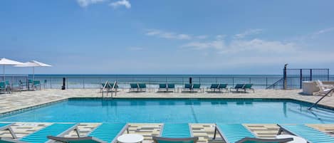 Un espace piscine avec des chaises longues et des parasols, un ciel dégagé et une vue sur l’océan.