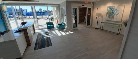 Un hall d’entrée spacieux avec un comptoir de réception, deux fauteuils turquoise, une grande fenêtre donnant sur l’extérieur et un tableau accroché au mur.