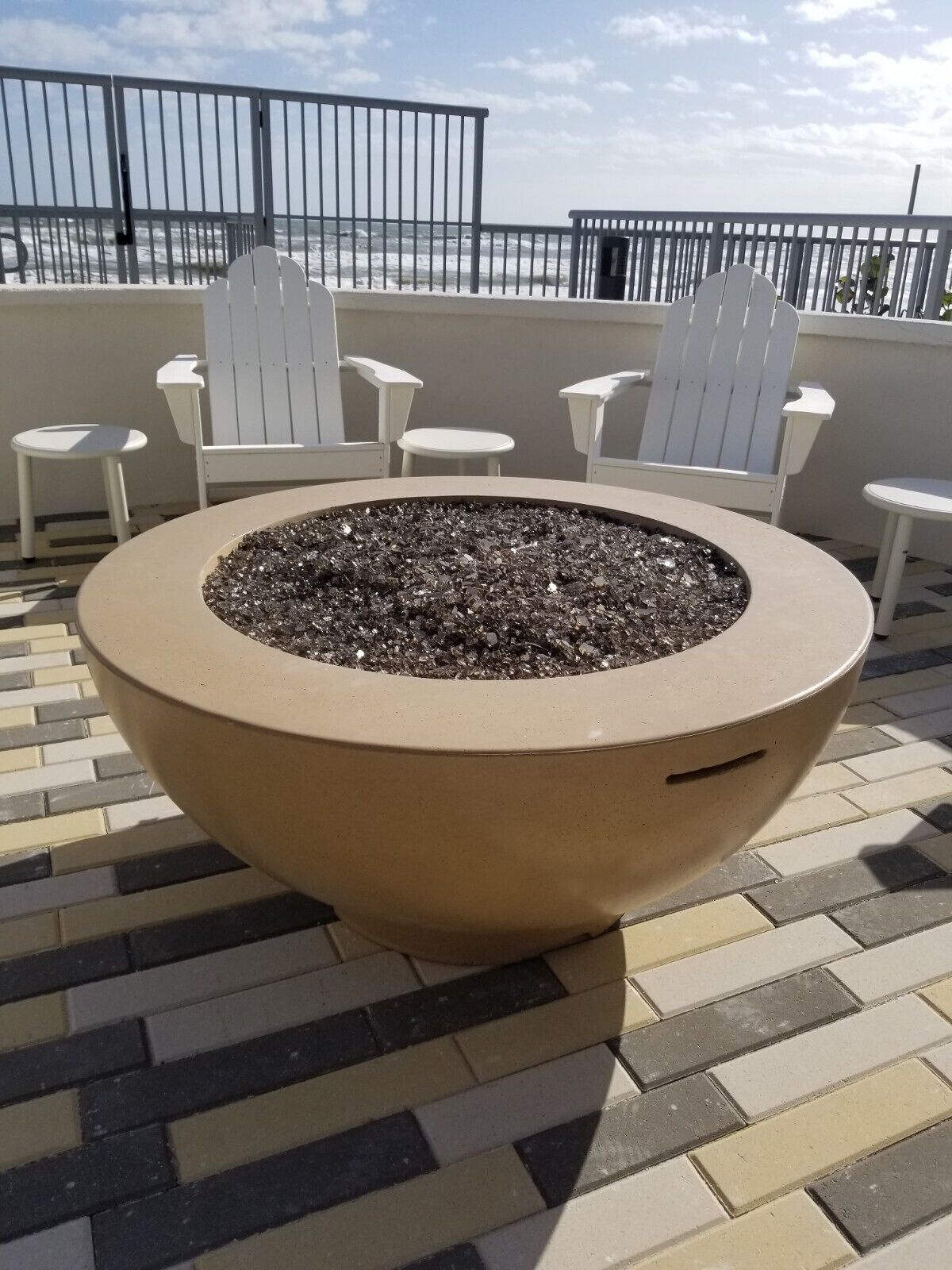 Un espacio de estar circular al aire libre con una chimenea, rodeado de sillas y mesas blancas, en un patio empedrado con vistas al océano.