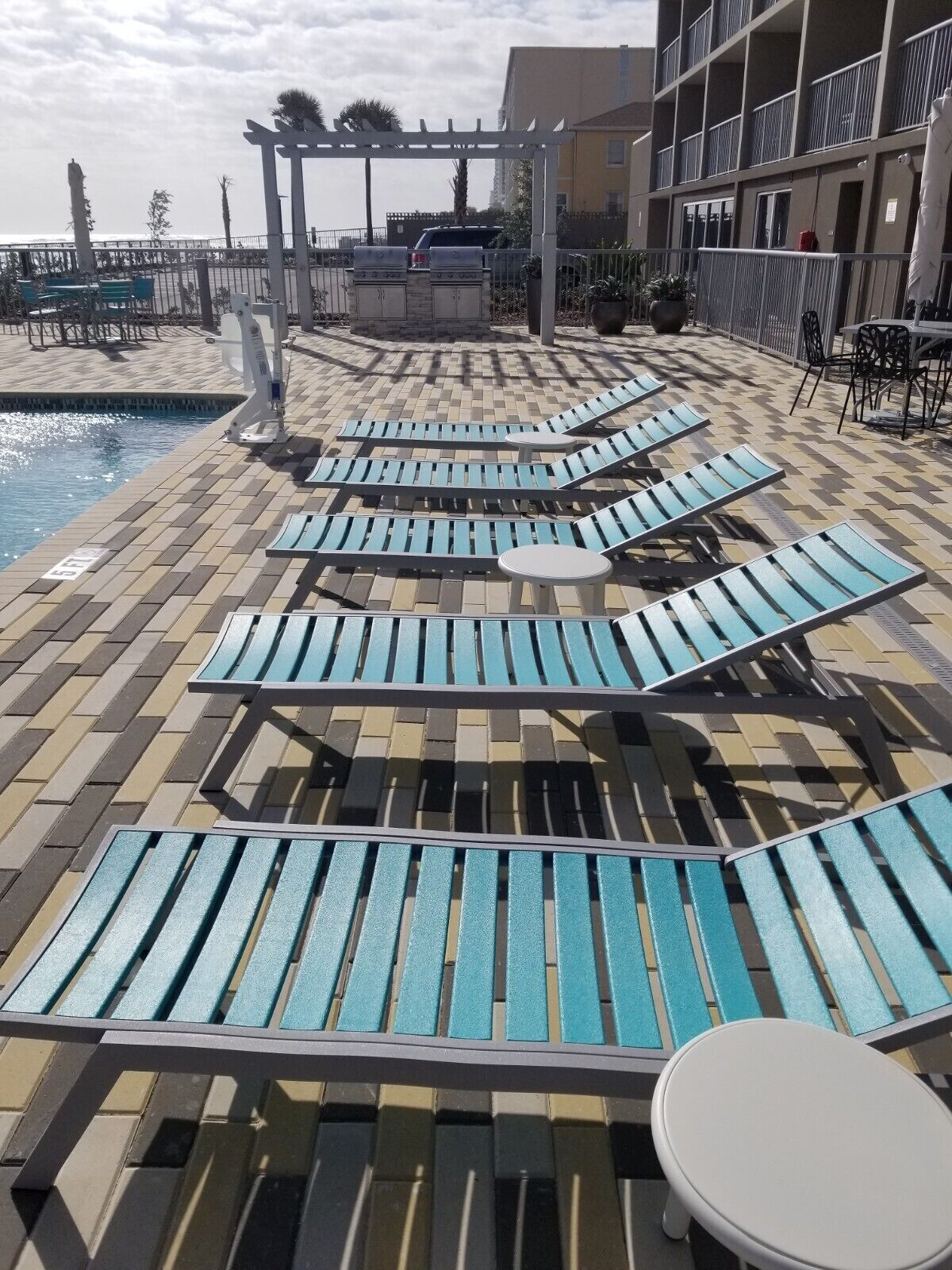 A poolside area with lounge chairs and a small table, a pergola, and a building in the background.