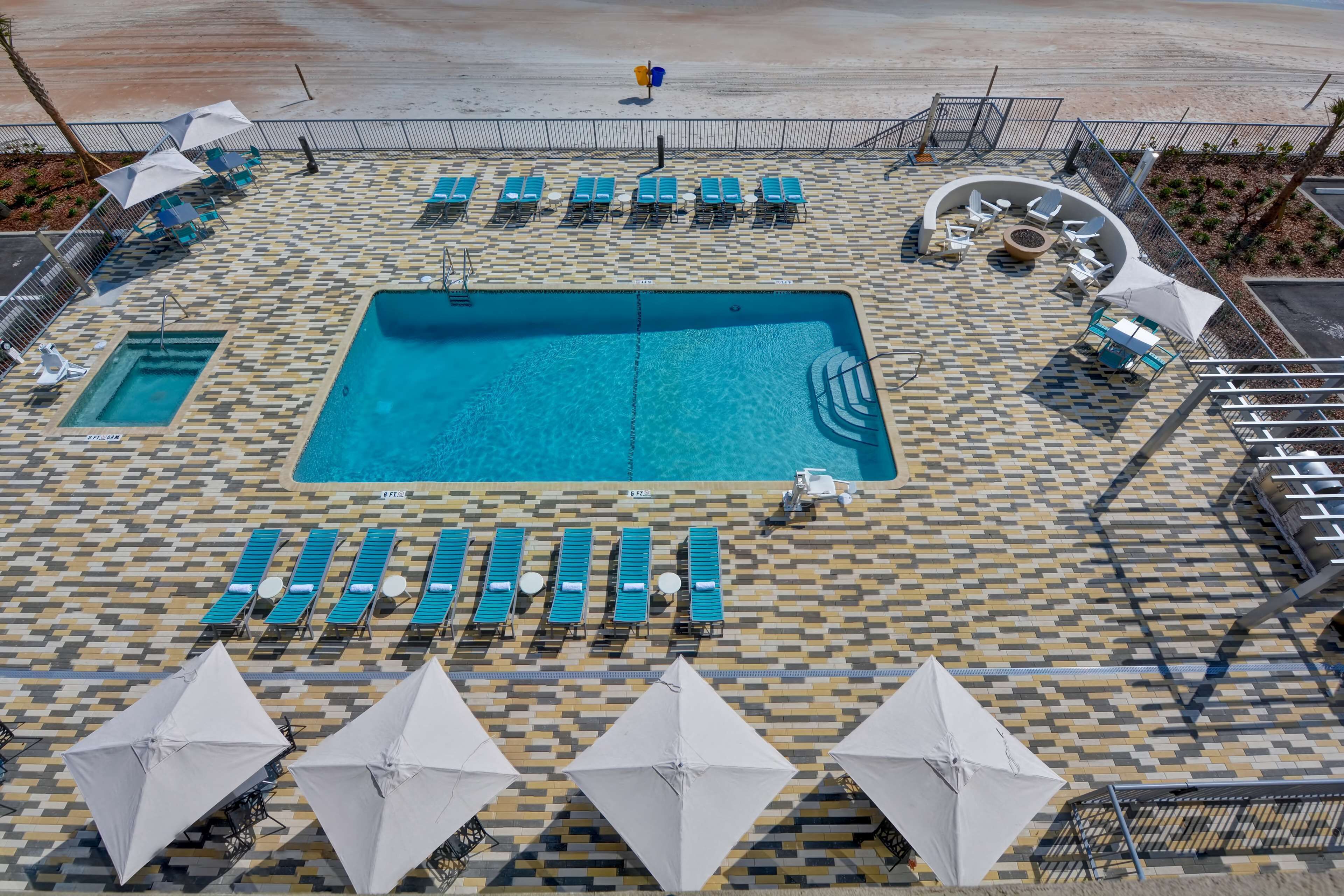 Zona de piscina con sillas reclinables azules y sombrillas, piscina central y suelo estampado.