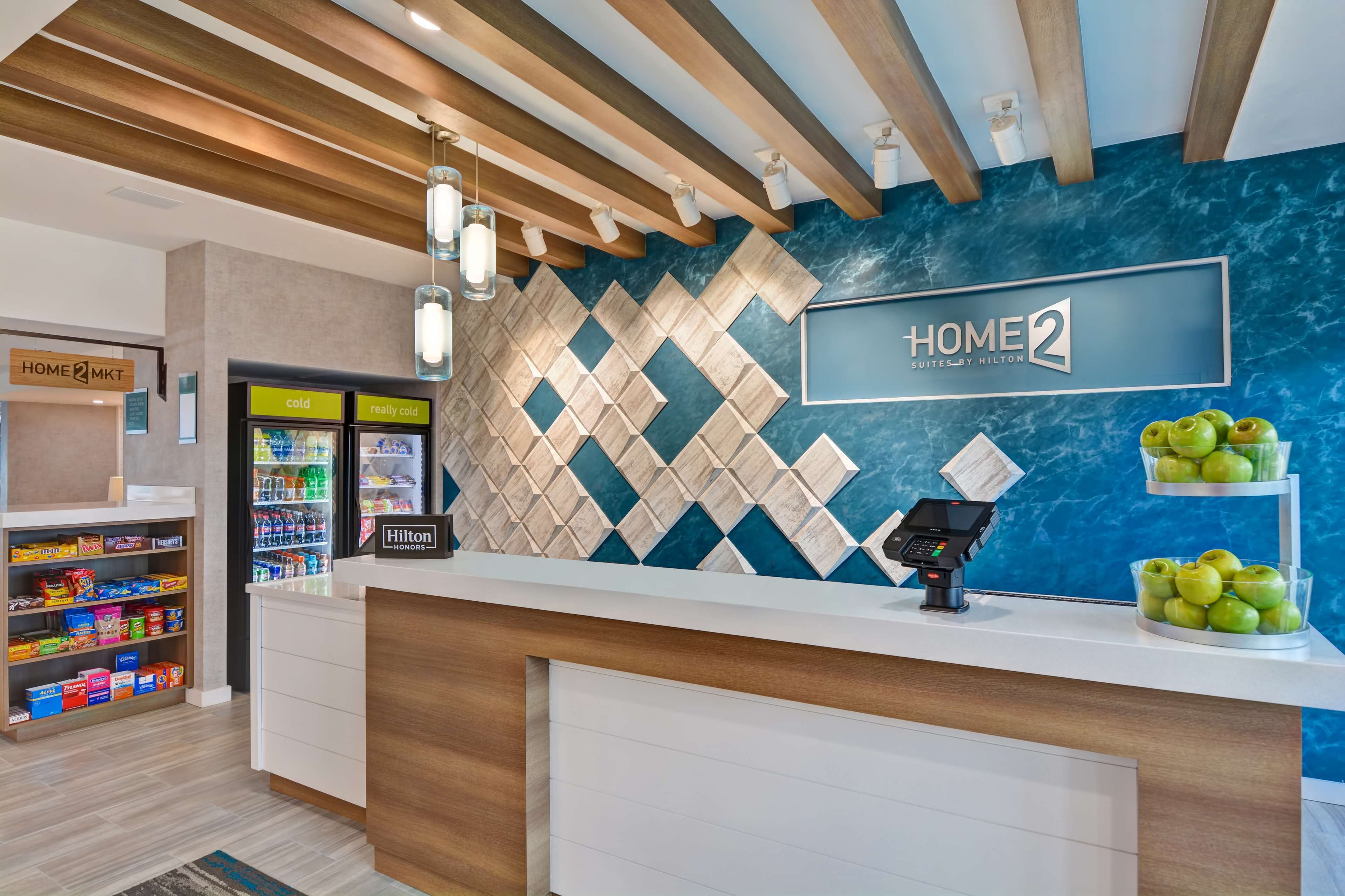 A hotel reception area with a modern design, featuring a marble wall, a digital kiosk, and a variety of snacks and beverages available.