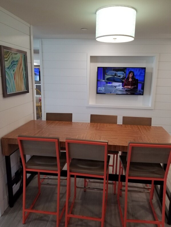 A dining area with a wooden table and red chairs, a television mounted on the wall, and a painting on the wall.