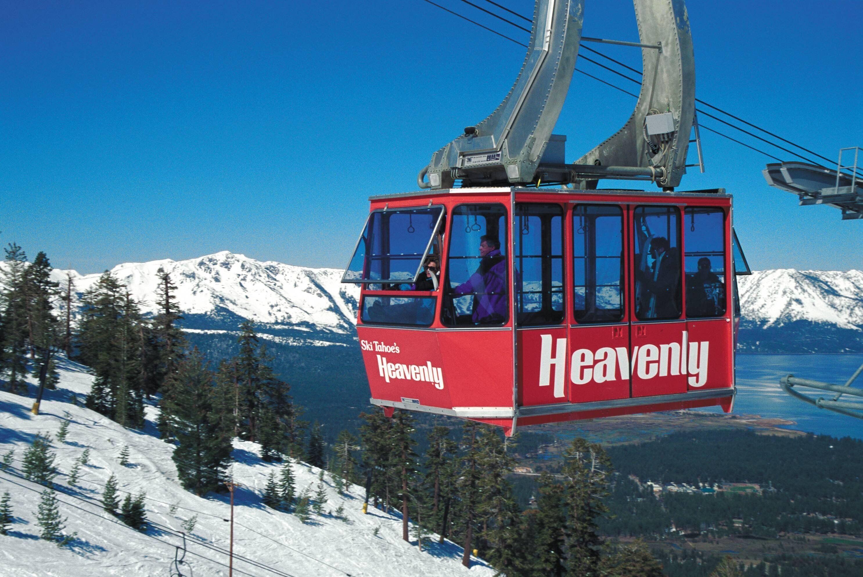 Une télécabine rouge sur laquelle on peut lire le mot « Heavenly », suspendue à des câbles, avec en arrière-plan des montagnes enneigées et un ciel bleu dégagé.