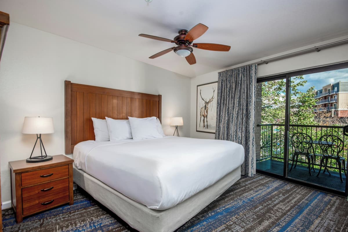 A hotel room with a large bed, a wooden headboard, two bedside tables with lamps, a ceiling fan, and a balcony with outdoor furniture.