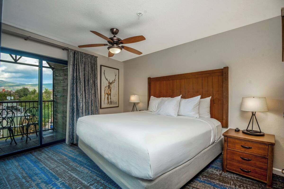 A hotel room with a large bed, a ceiling fan, a nightstand with a lamp, a view of the outdoors through a glass door, and a balcony with outdoor furniture.