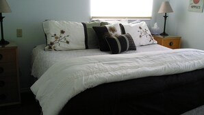 A neatly made bed with a white and black bedspread, multiple pillows, and a lace-trimmed footboard. There are two bedside tables with lamps and a small wooden nightstand with a lamp.