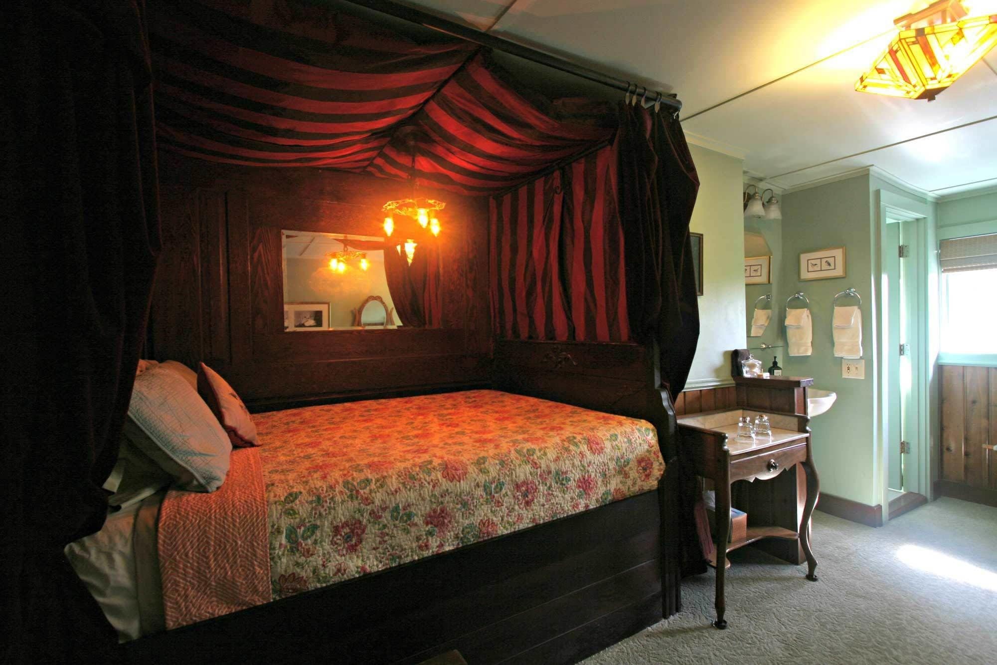 Uma cama de dossel tradicional com colcha florida e cortinas escuras. Um criado-mudo de madeira com uma lâmpada e um espelho fica ao lado da cama. O quarto tem uma parede verde clara e uma janela com vista para o exterior.