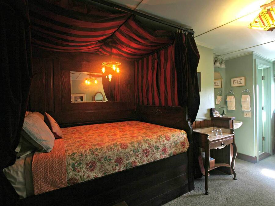 Uma cama de dossel tradicional com colcha florida e cortinas escuras. Um criado-mudo de madeira com uma lâmpada e um espelho fica ao lado da cama. O quarto tem uma parede verde clara e uma janela com vista para o exterior.