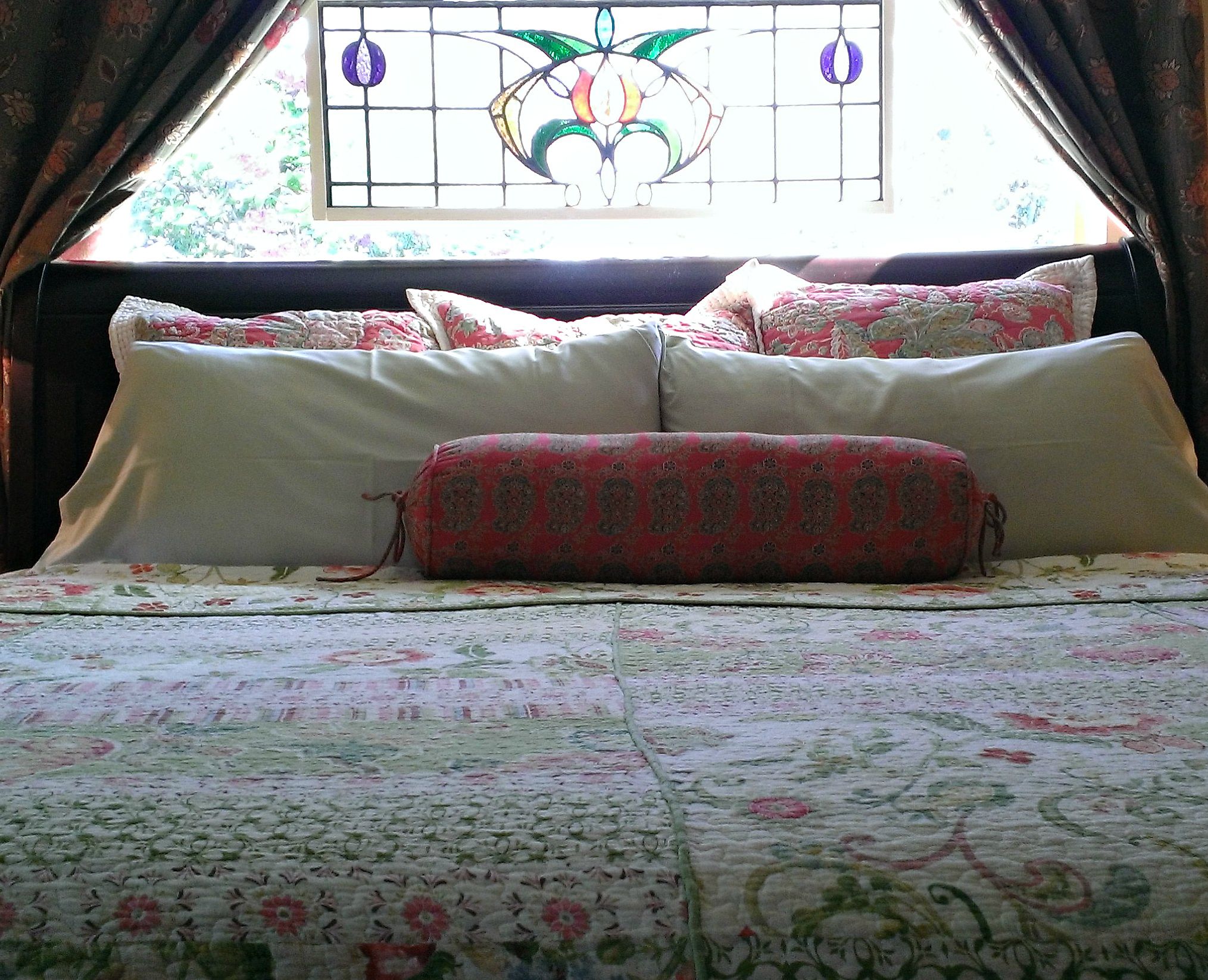 Una cama con ropa de cama floral y un cojín estampado, una ventana con un diseño de vitral y cortinas.