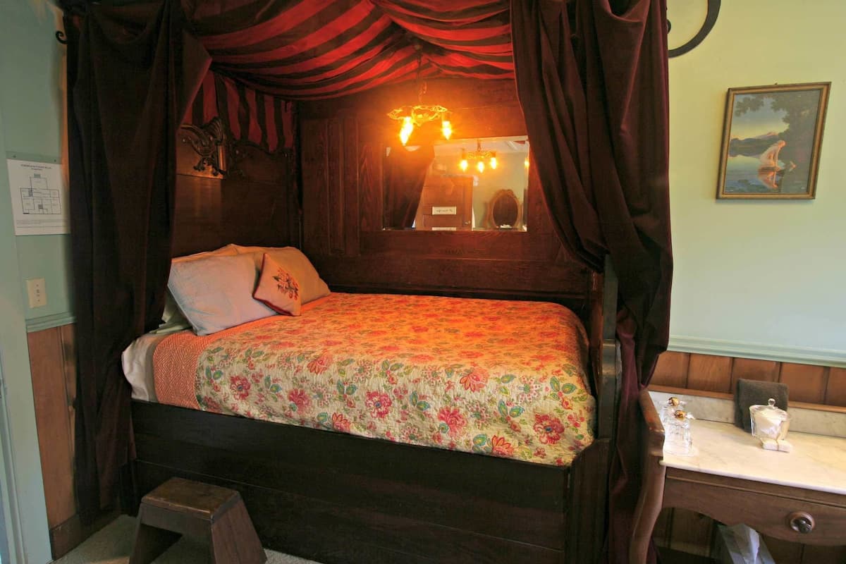 A bed with a floral bedspread, a nightstand with a mirror and a small lamp, and a painting on the wall.