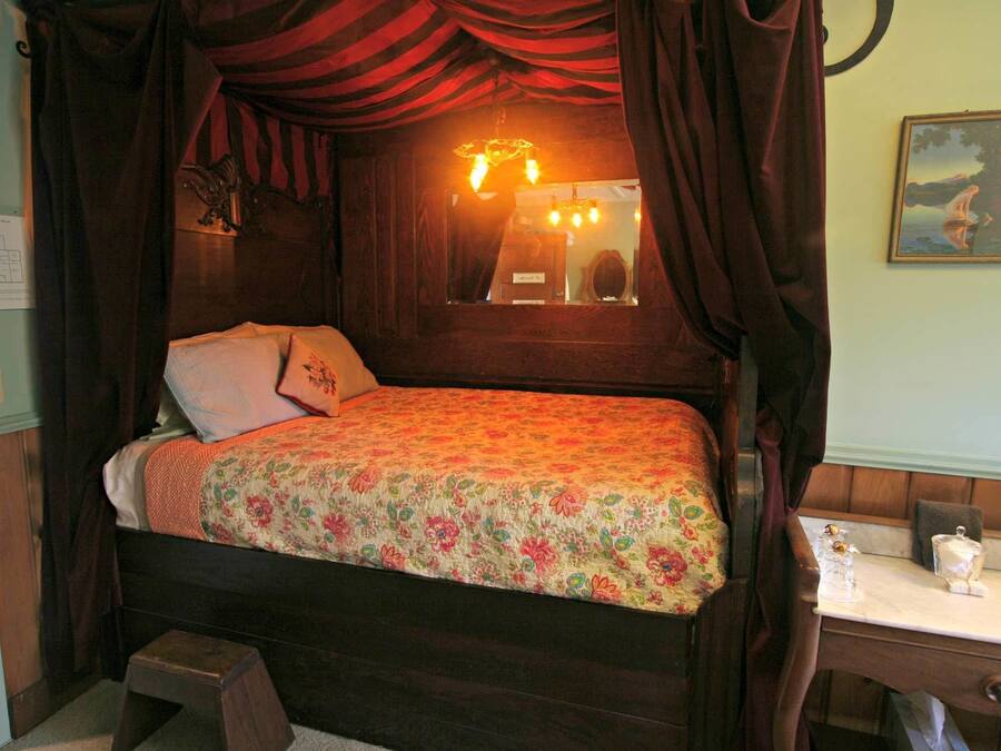 Una cama con colcha estampada, una mesita de noche con espejo y una lámpara pequeña, y un cuadro en la pared.