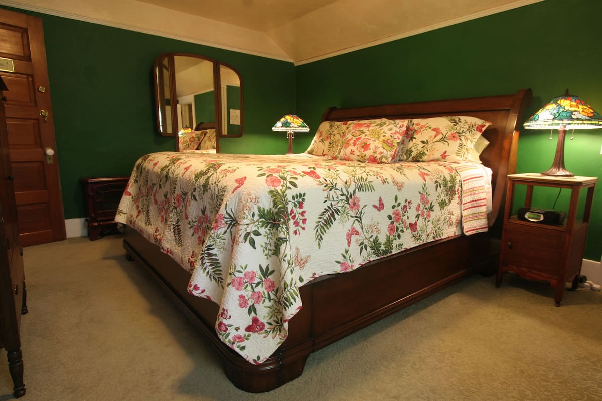 A bedroom with a floral bedspread, a wooden headboard, a nightstand with a lamp, and a mirror on the wall.