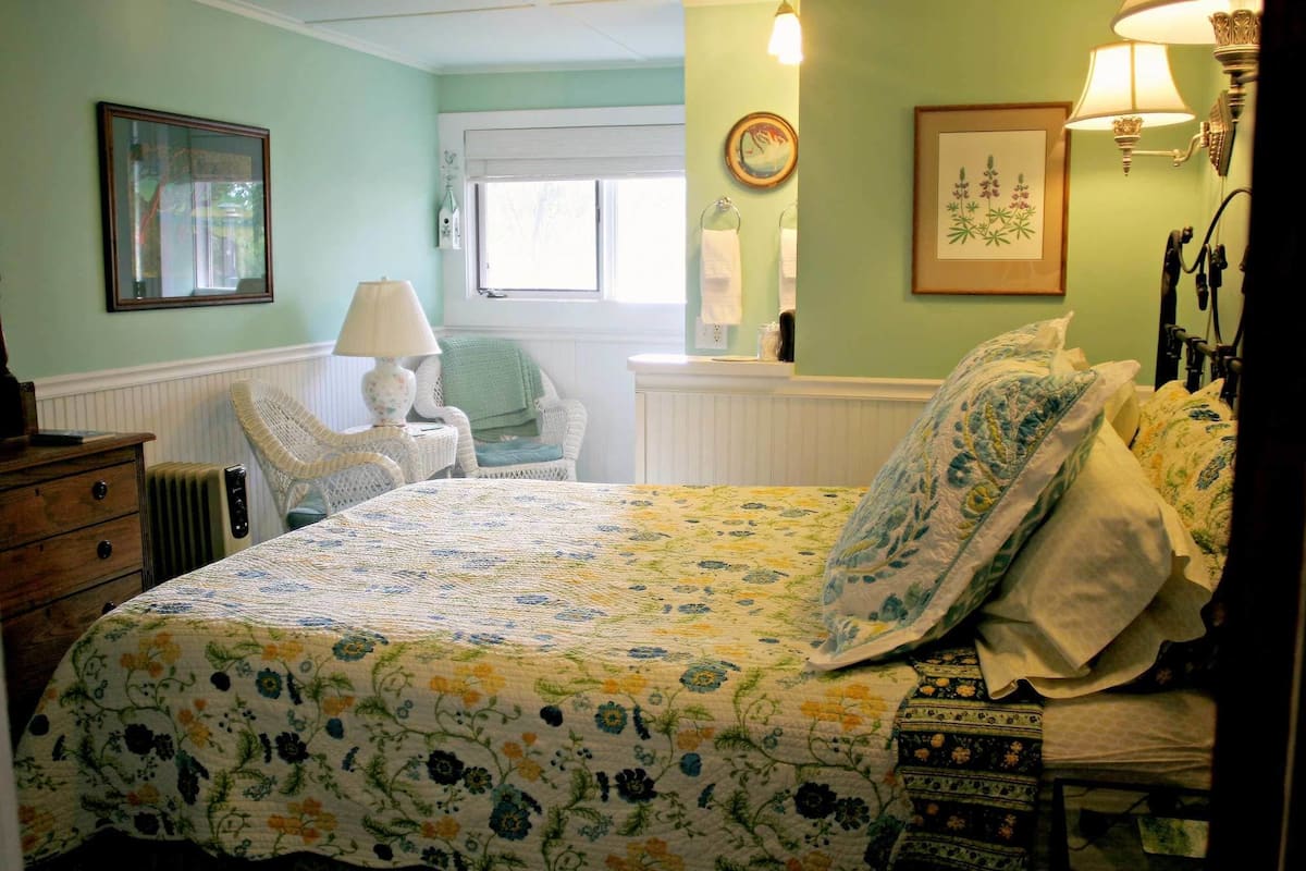 A bedroom with a floral bedspread, a wicker chair, a bedside table with a lamp, and a dresser.