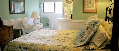 Une chambre à coucher avec une couvre-lit à motifs floraux, une chaise en osier, une table de chevet avec une lampe et une commode.