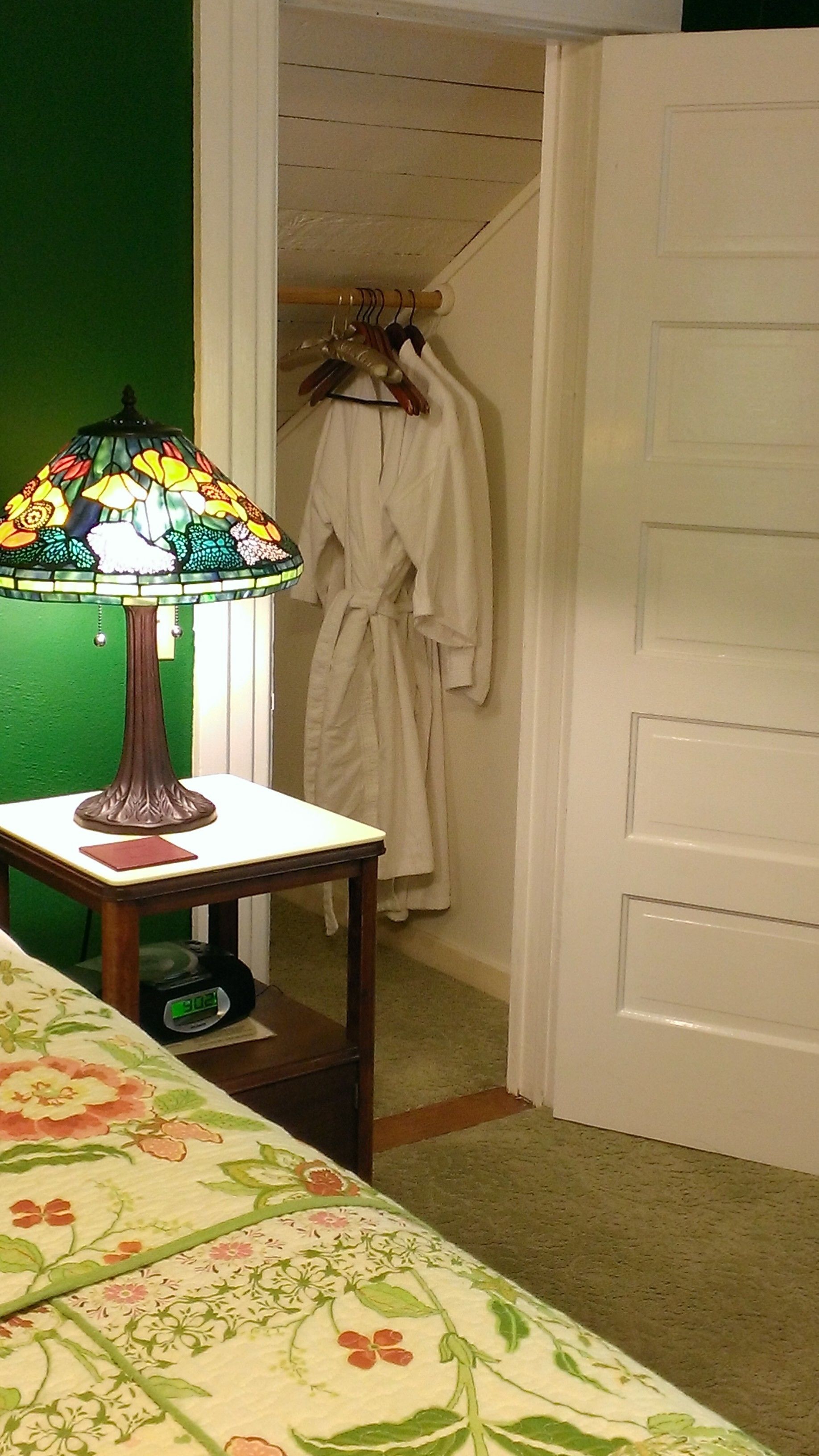 A room with a bed, a table with a lamp, a green wall, a white door, and a robe hanging on a rack.
