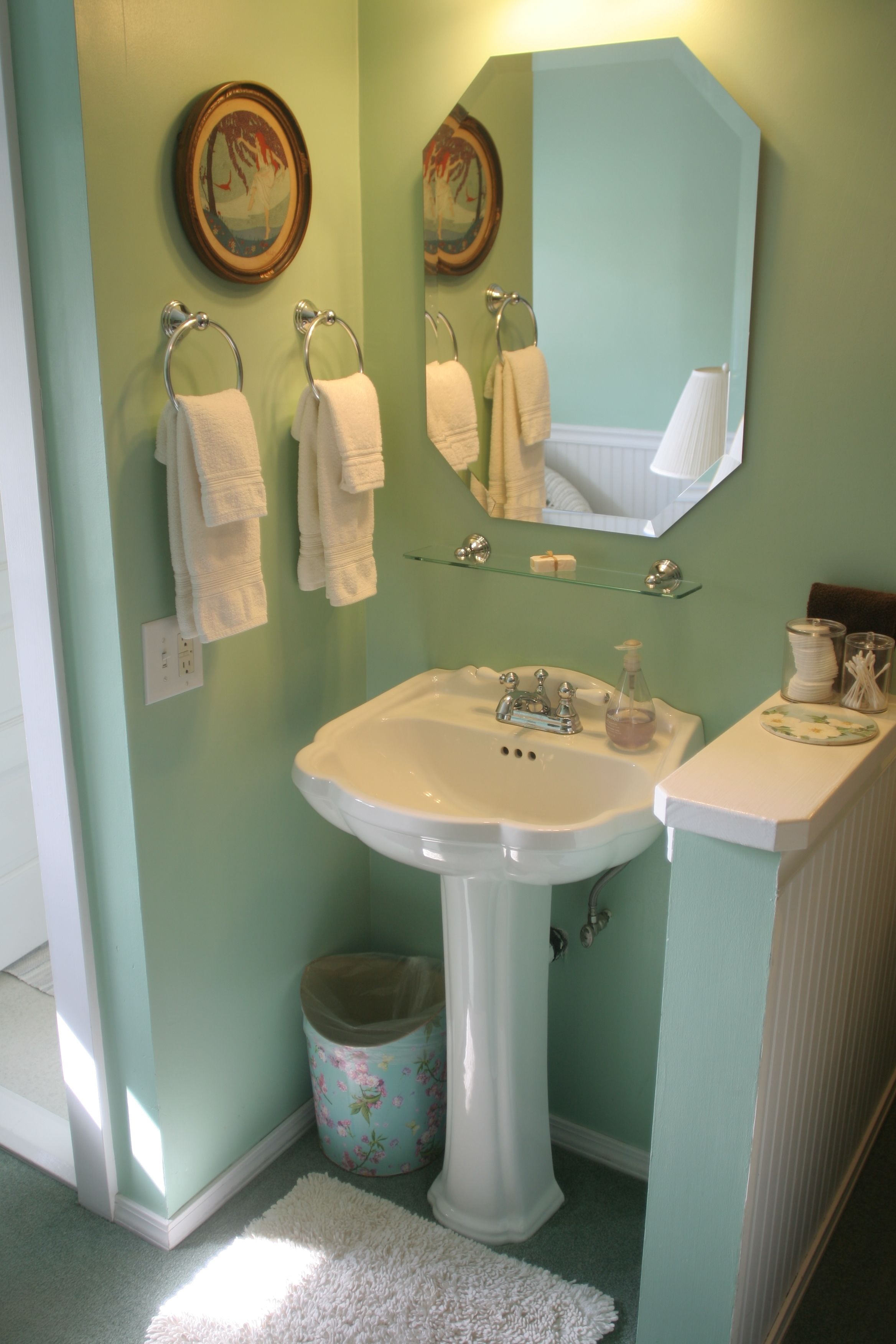 A bathroom with a pedestal sink, two wall-mounted towel rings, a mirror, and a small trash bin.