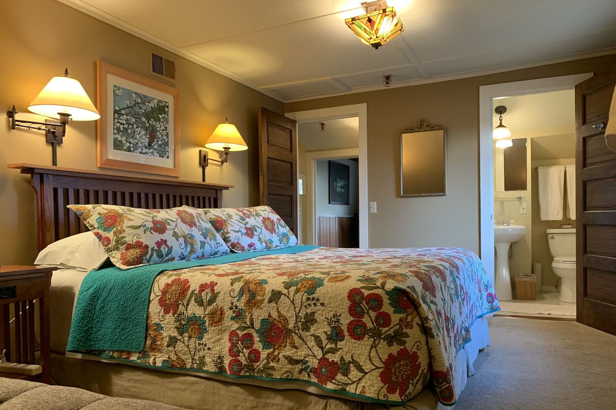 A bedroom with a large bed, two wall-mounted lamps, a framed floral painting, and a view into the bathroom.