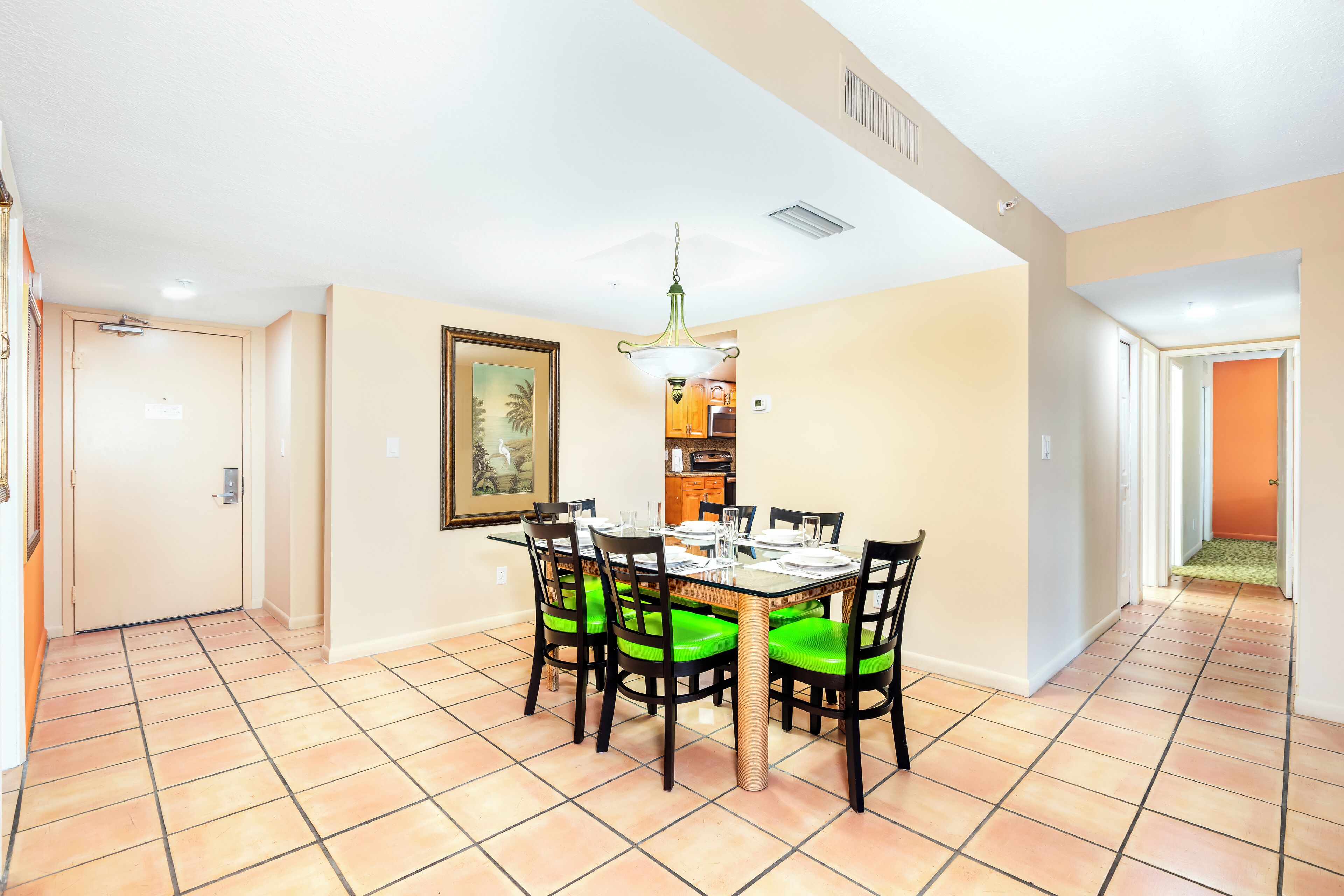 Un coin repas avec une table dressée pour six personnes, entourée de chaises dotées de coussins verts, un luminaire suspendu et un tableau encadré au mur.