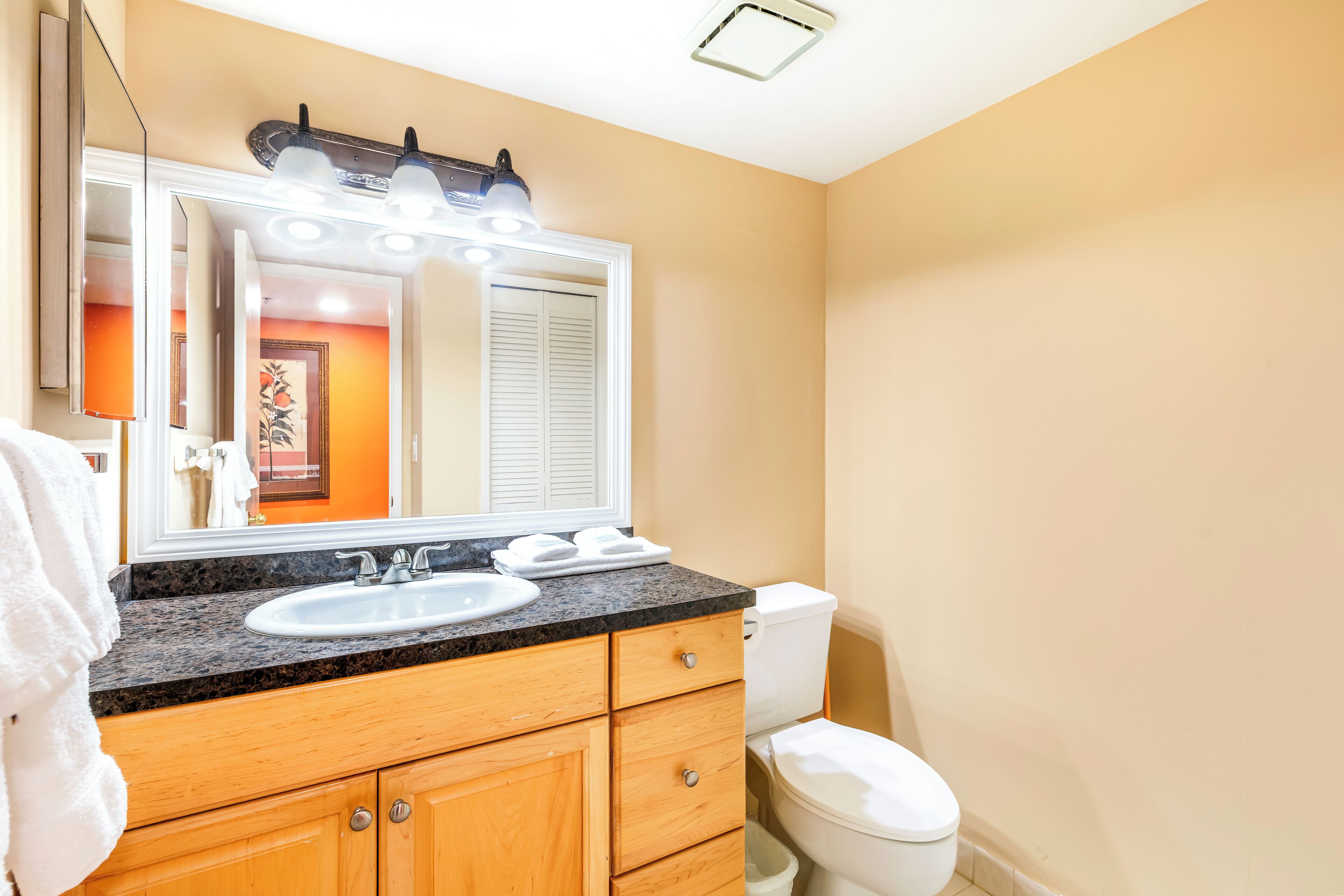A bathroom with a granite countertop sink, wooden cabinets, a white toilet, and a large mirror with lights.