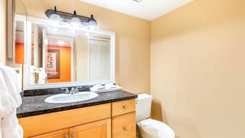 Un baño con lavamanos de encimera de granito, armarios de madera, inodoro blanco y un espejo grande con iluminación.