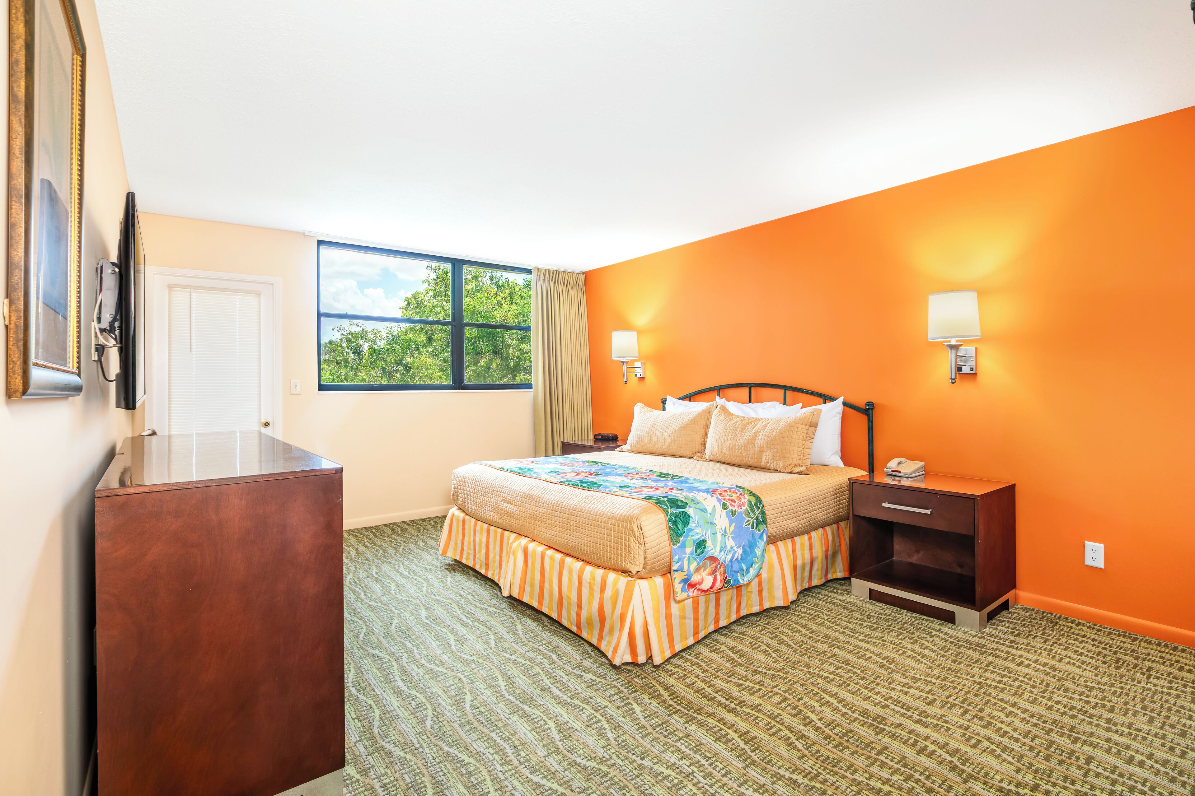 A hotel room with a bed, a nightstand, a desk, and a window with a view of greenery.