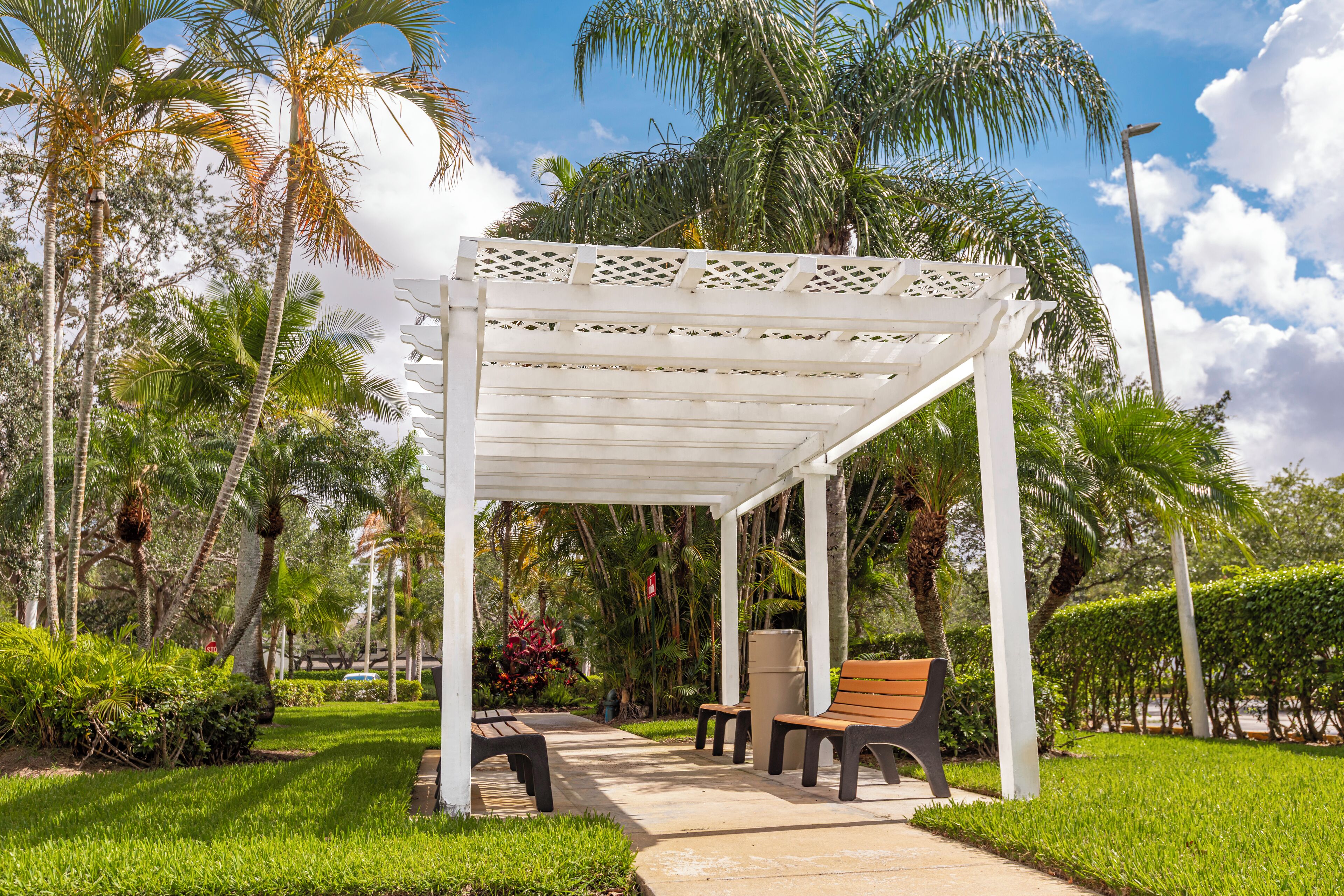 A covered outdoor seating area with a white pergola, benches, and a small table, surrounded by lush greenery and palm trees.