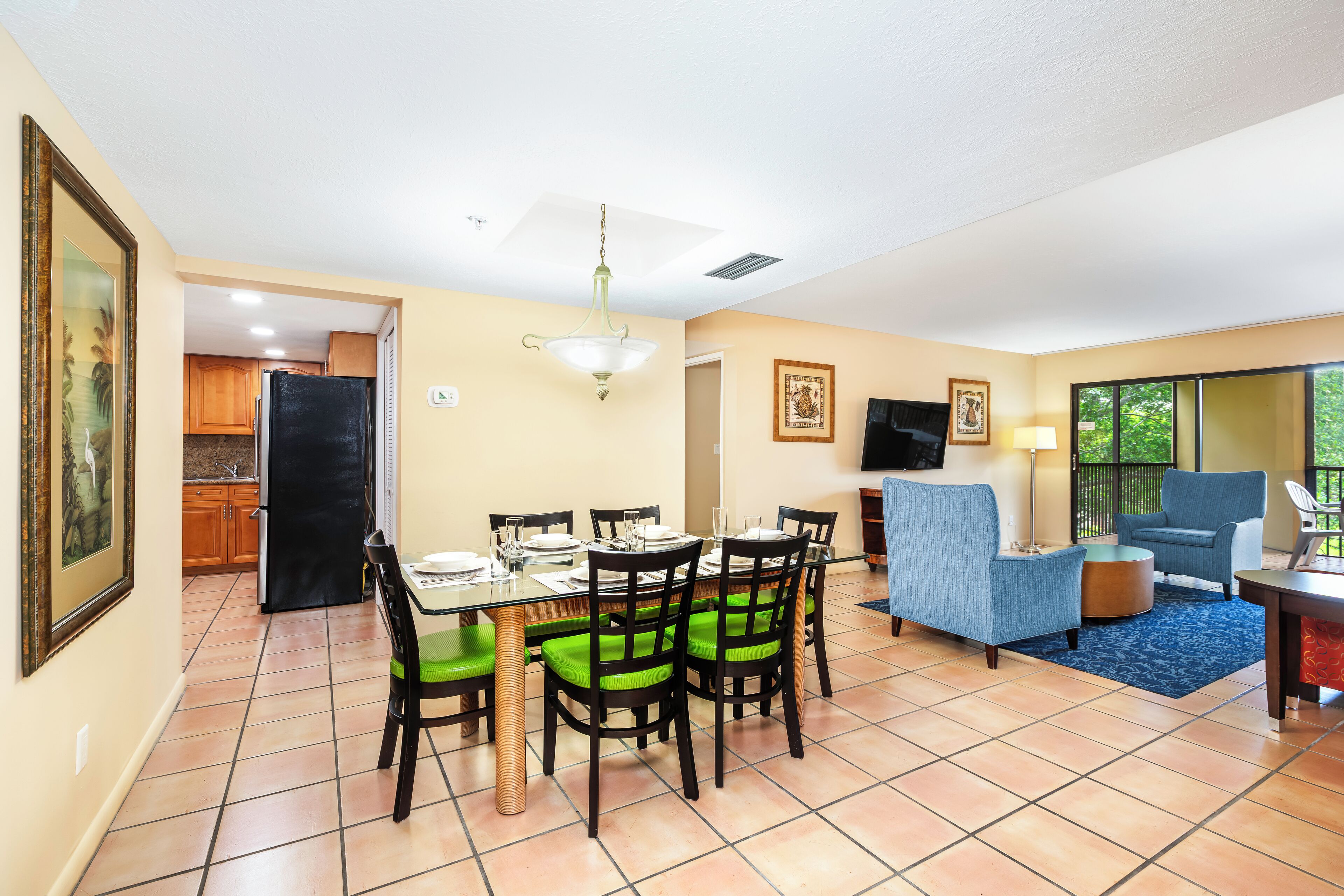 Un coin repas avec une table en verre, des chaises à coussins verts et un réfrigérateur noir.