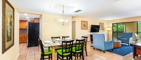Uma sala de jantar com mesa de vidro, cadeiras estofadas verdes e uma geladeira preta.