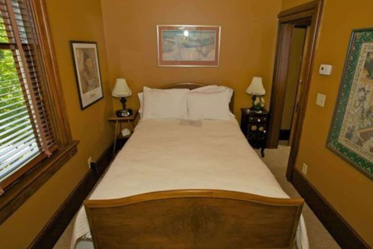 A bedroom with a wooden bed, two bedside tables with lamps, a framed picture on the wall, and a window with blinds.