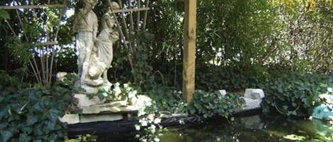 Un jardin agrémenté d’une statue représentant deux personnages, d’un bassin et d’une végétation luxuriante.
